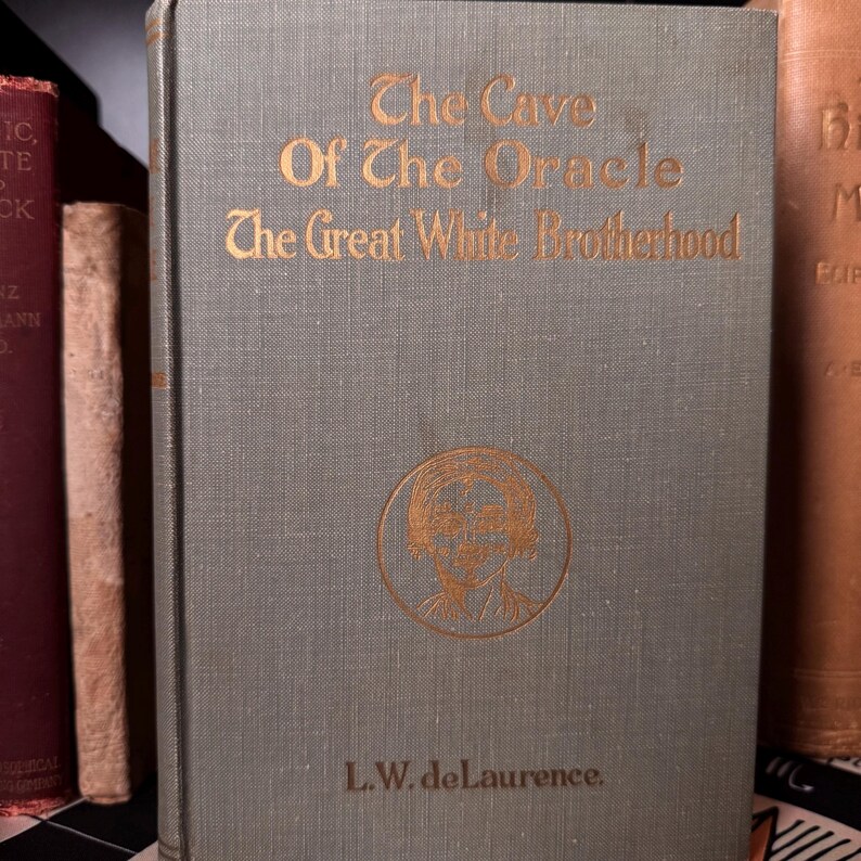 May include: A vintage book titled "The Cave Of The Oracle The Great White Brotherhood" with gold lettering on a gray-green cover. The cover features a circular illustration and the author's name, L.W. de Laurence. Other books are visible in the background.