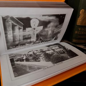 May include: An open book with black and white photographs of ancient ruins. The top image shows an Ankh symbol and the text "A Collage Of Ancient Ruins On Tama-Re". The book has an orange cover and is next to a green book.