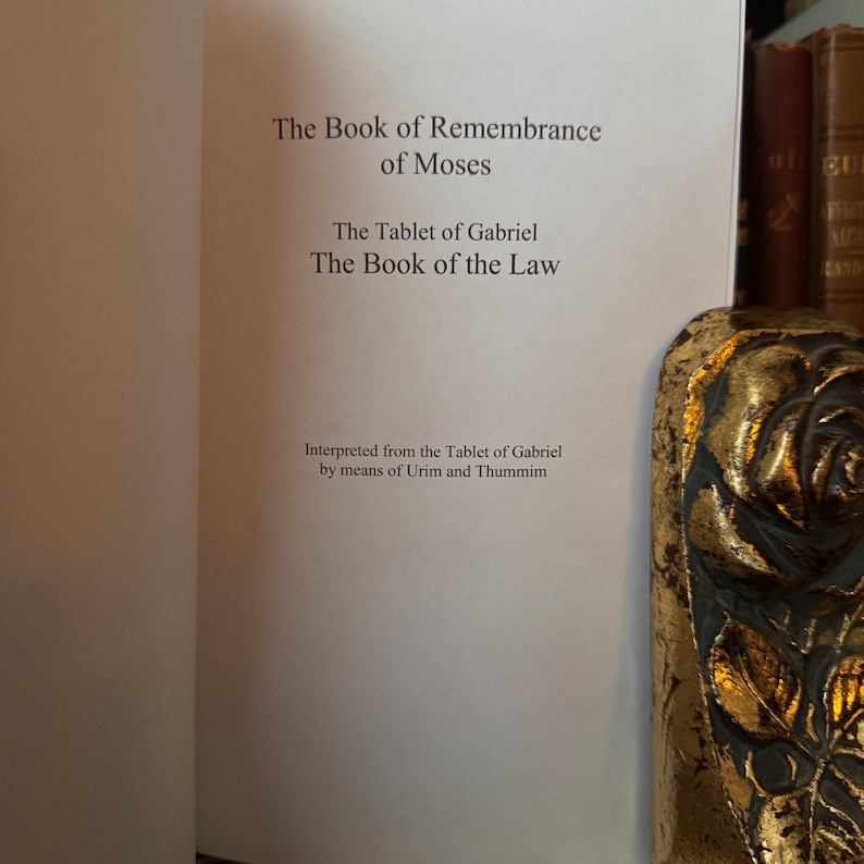 Peut inclure: Un livre ouvert avec le texte "The Book of Remembrance of Moses", "The Tablet of Gabriel" et "The Book of the Law". Le texte "Interpr&eacute;t&eacute; de la Tablette de Gabriel par le biais d'Urim et Thummim" est &eacute;galement visible. Un objet d&eacute;coratif dor&eacute; est en arri&egrave;re-plan.