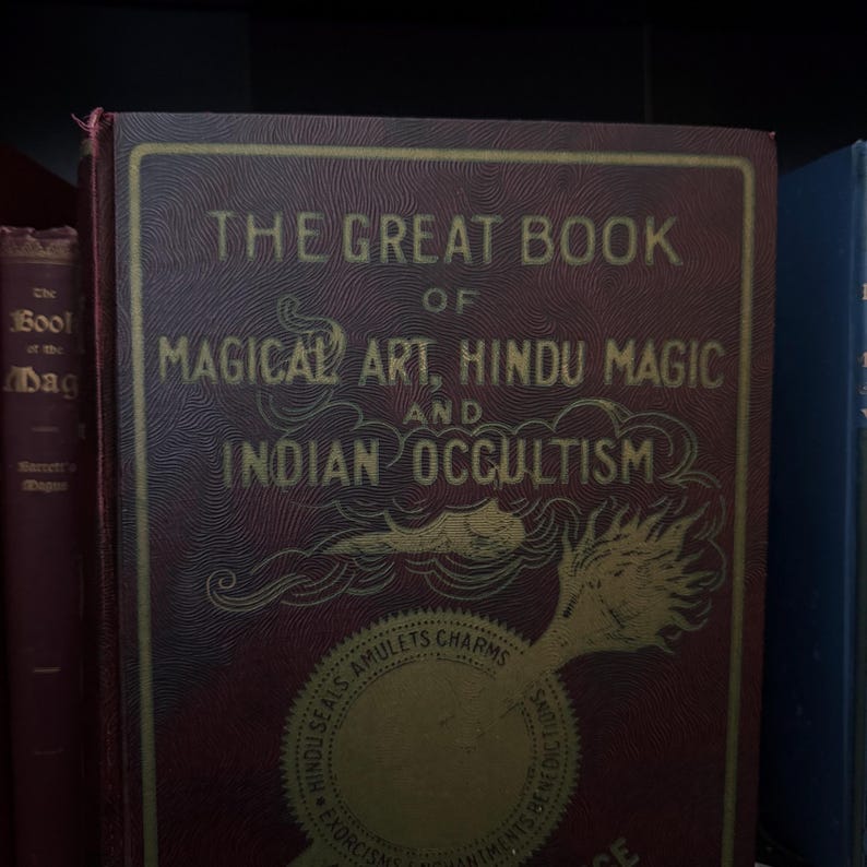 Pu&ograve; includere: Un libro color borgogna con lettere dorate intitolato "The Great Book of Magical Art, Hindu Magic and Indian Occultism". La copertina presenta un'illustrazione dorata di una mano che tiene una bacchetta con una nuvola e fiamme.