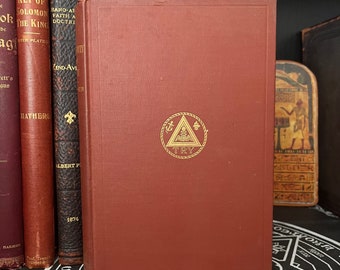 Puede incluir: Un libro de tapa dura rojo con letras doradas y un emblema dorado en la portada. El emblema es un triángulo con una calavera y huesos cruzados en su interior, rodeado por un diseño de cuerda. El texto en la portada dice "TRY".