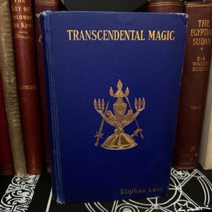 May include: A blue hardback book titled "Transcendental Magic" by Eliphas Levi. The book cover features a gold illustration of a candelabra with swords and a helmet.