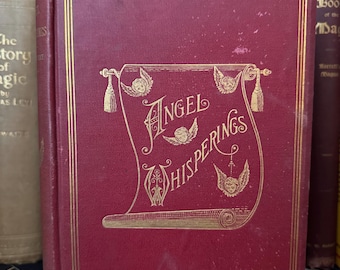 Może przedstawiać: Stara książka z bordową okładką i złotymi literami. Tytuł "Angel Whisperings" jest wyświetlany na ozdobnym zwoju z ilustracjami aniołów. Książka ma złote obramowanie i jest częścią kolekcji.