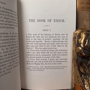 May include: Open book with the title "THE BOOK OF ENOCH" on the right page. The text is in black font on a cream-colored page. A decorative gold-colored object is on the right side of the image. The left page has text as well.