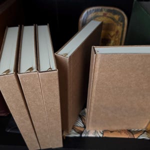 May include: A collection of hardcover books with kraft paper covers and cream-colored pages. The books are arranged in a shelf, with some standing upright and others leaning. The spines are plain, and the overall aesthetic is minimalist and natural.