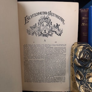 Puede incluir: P&aacute;gina de un libro antiguo con el t&iacute;tulo "Encyclopaedia Freemasonry" en letras ornamentadas. La p&aacute;gina presenta texto y un emblema detallado. Un objeto decorativo con detalles dorados y azules es visible a la derecha.