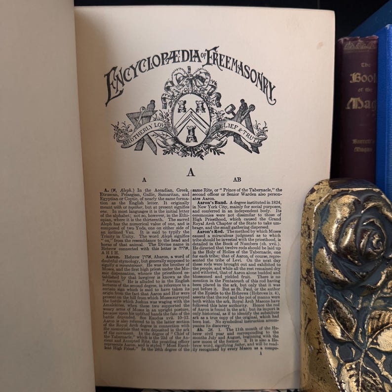 Puede incluir: P&aacute;gina de un libro antiguo con el t&iacute;tulo "Encyclopaedia Freemasonry" en letras ornamentadas. La p&aacute;gina presenta texto y un emblema detallado. Un objeto decorativo con detalles dorados y azules es visible a la derecha.