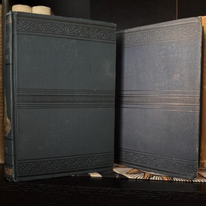 May include: Two antique books with dark blue covers. The covers feature decorative black line designs at the top, middle, and bottom. The books are displayed on a dark surface, suggesting a vintage or historical theme.