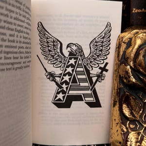 May include: A black and white illustration of an eagle with wings spread, holding two crossed rifles. The eagle is perched on top of a large letter "A" with five stars and stripes. The letter "A" is surrounded by five stars.