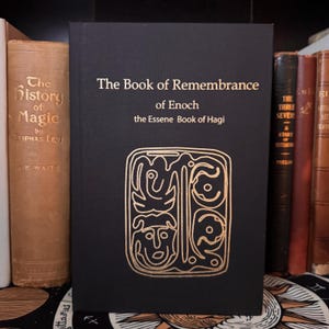May include: A black book titled "The Book of Remembrance of Enoch" with gold lettering and an ornate gold design. The book is standing upright on a shelf with other antique books. The book's cover features the text "the Essene Book of Hagi".