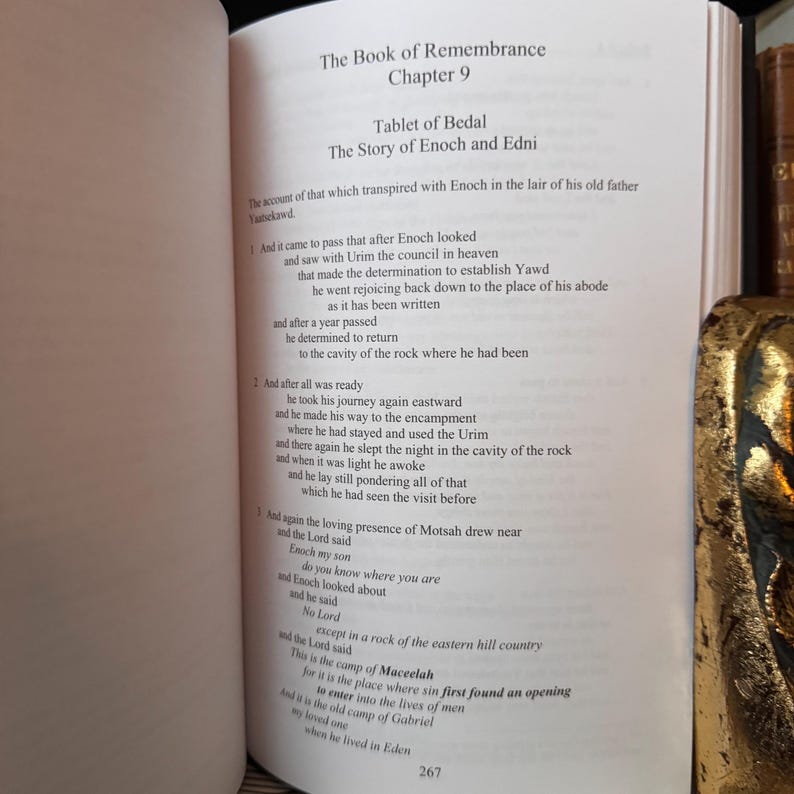 May include: Open book with text titled "The Book of Remembrance Chapter 9". The text describes "Tablet of Bedal The Story of Enoch and Edni". The page number "267" is visible at the bottom. A gold-colored object is visible on the right side.