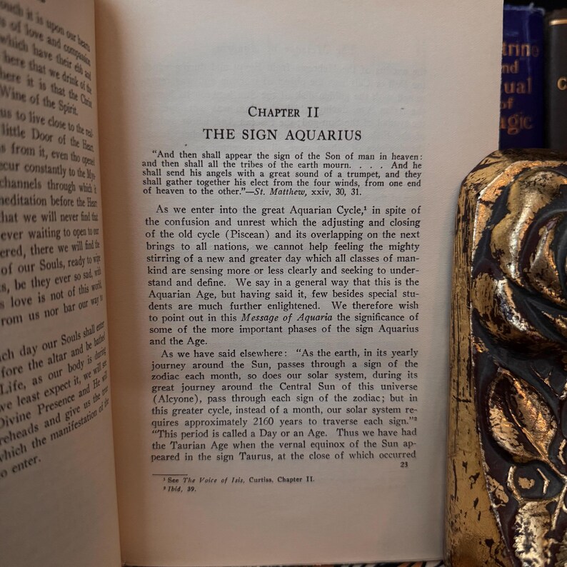 May include: A vintage book with a yellowed page open to Chapter II, titled "The Sign Aquarius." The text discusses the astrological sign Aquarius and its significance in the Aquarian Age.