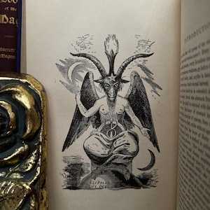 May include: A black and white illustration of a goat-like figure with horns and wings. The figure is sitting on a throne with a pentagram on its head. The text "CLIPPAS LEVI DEL" is written below the figure.
