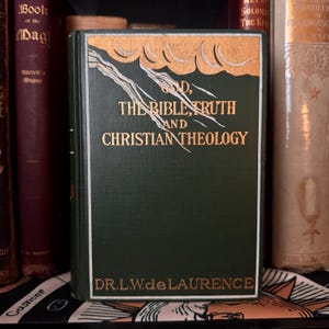 May include: A dark green book with the title "God, The Bible, Truth and Christian Theology" in gold lettering. The author's name, Dr. L.W. de Laurence, is printed at the bottom. The book is displayed amongst other antique books.