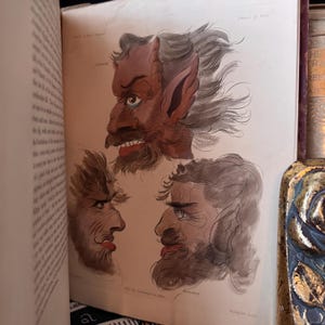 Puede incluir: Un libro abierto muestra tres ilustraciones de rostros demon&iacute;acos. Los dibujos son detallados, con tonos rojos, marrones y grises. La ilustraci&oacute;n superior presenta orejas grandes y una barba. Las p&aacute;ginas del libro tienen un aspecto envejecido.