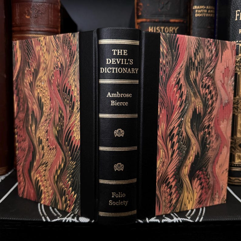 May include: A black leather-bound book with gold lettering titled "The Devil's Dictionary" by Ambrose Bierce. The book is part of the Folio Society collection and has a marbled cover design.