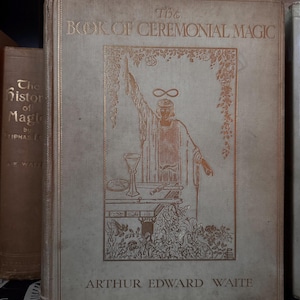 May include: A vintage book titled "The Book of Ceremonial Magic" by Arthur Edward Waite. The cover features a gold illustration of a robed figure with an infinity symbol above their head. The book is light beige with gold lettering.