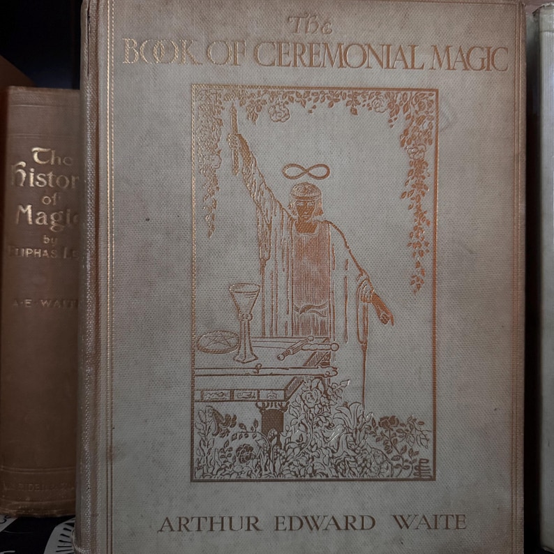 May include: A vintage book titled "The Book of Ceremonial Magic" by Arthur Edward Waite. The cover features a gold illustration of a robed figure with an infinity symbol above their head. The book is light beige with gold lettering.