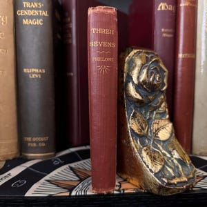 May include: A red hardcover book titled "Three Sevens" by Phelons, with a gold sunburst design on the cover, is being held upright by a gold-painted metal bookend shaped like a rose.