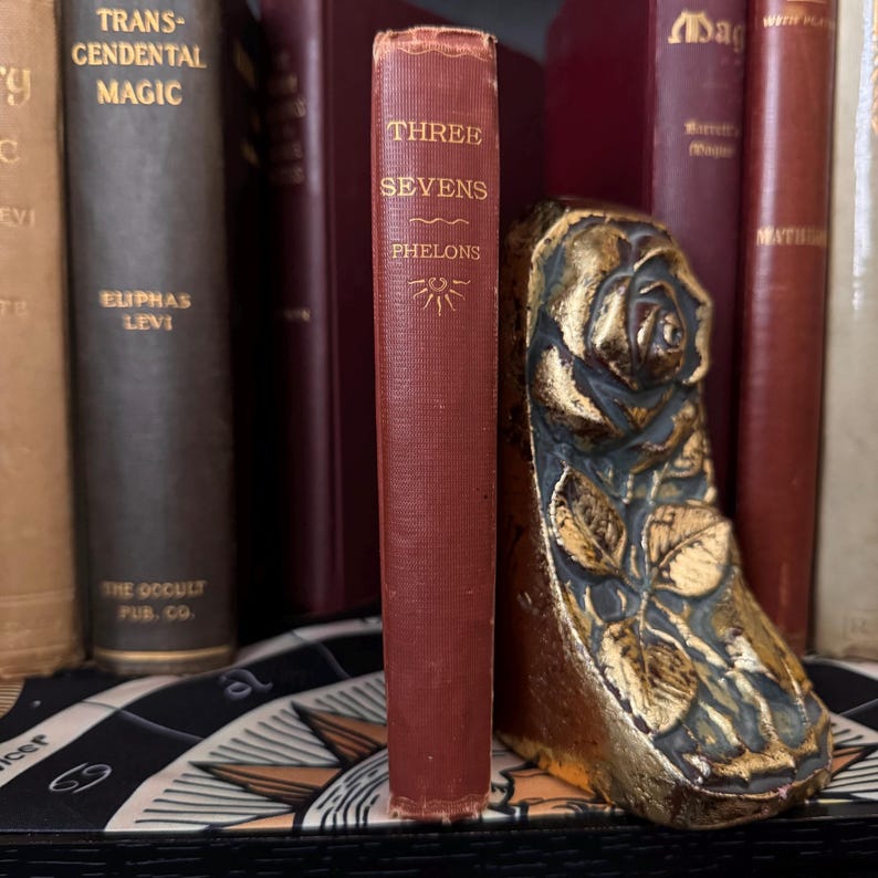 May include: A red hardcover book titled "Three Sevens" by Phelons, with a gold sunburst design on the cover, is being held upright by a gold-painted metal bookend shaped like a rose.