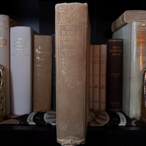 May include: A vintage book titled "The Book of Ceremonial Magic" by Arthur Edward Waite. The book has a beige cover with gold lettering and decorative floral patterns. Other antique books are visible on a shelf.