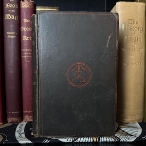 May include: A dark brown hardcover book featuring a circular red emblem on the front cover. The book is positioned between other vintage books with visible titles including The Book of the Magus and The History of Magic, all standing upright.