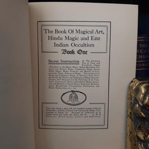 Pu&ograve; includere: Un libro vintage intitolato "The Book of Magical Art, Hindu Magic and East Indian Occultism" con il sottotitolo "Secret Instruction for the Exclusive Use of True and Faithful Chelas (Disciples)". La copertina del libro &egrave; bianca con testo nero e una piccola illustrazione di una persona seduta.