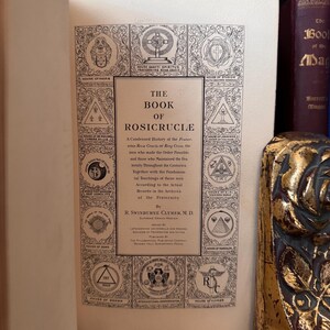 Może przedstawiać: Antyczna książka zatytułowana "The Book of Rosicrucia" z kr&oacute;tką historią Zakonu R&oacute;żokrzyżowc&oacute;w. Okładka zawiera ozdobny wz&oacute;r z symbolami i tekstem.