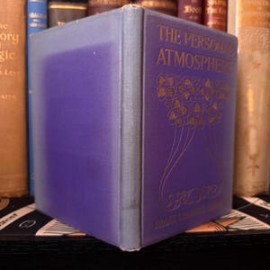 Puede incluir: Un libro antiguo titulado "THE PERSONAL ATMOSPHERE" con una cubierta morada y letras doradas. El lomo del libro es de un tono morado m&aacute;s claro. La portada presenta un dise&ntilde;o floral ornamentado. El libro se exhibe entre otros libros antiguos.