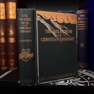 May include: A vintage hardback book titled "God, The Bible, Truth and Christian Theology" by Dr. L.W. de Laurence. The book cover is dark green with gold lettering and a decorative design of clouds and lightning bolts.