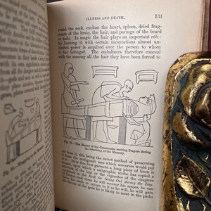 以下が含まれることがあります： 古代エジプトの儀式を描いた白黒のイラスト。祭司がテーブルに横たわる亡くなった人の上に儀式を行っている。祭司は長いローブと頭飾りをつけている。亡くなった人はリネン製の包帯で包まれている。このイラストは、古代エジプトの信仰と慣習に関する本からのものである。