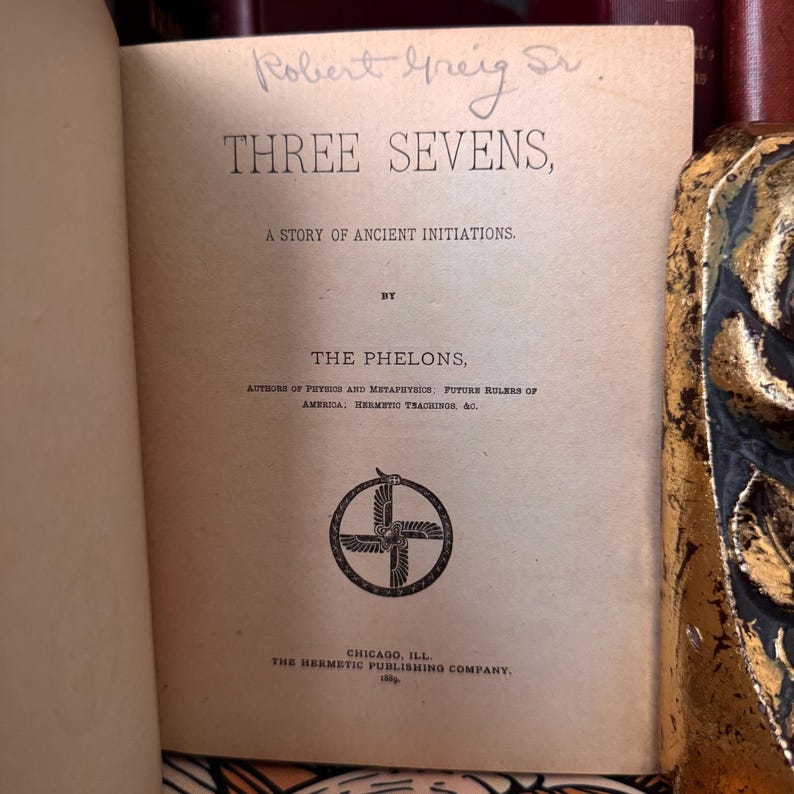May include: A vintage book titled "Three Sevens: A Story of Ancient Initiations" by The Phelons, authors of "Pythios and Metaphysics: Future Rulers of America: Hermetic Trachinor, 4c." The book cover features a black and white illustration of a cross with a winged serpent.