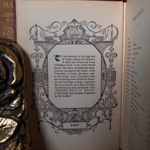 May include: An open book page with ornate black and white illustrations and text. The text includes the title "SACRED BOOK OF DEATH" and the year 1907. The page is from a vintage book with a gold-colored decorative edge.
