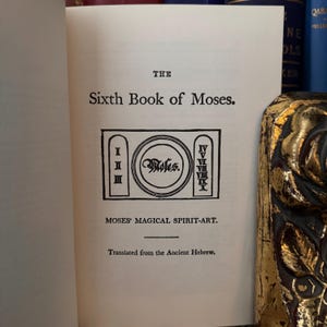 May include: A book titled "The Sixth Book of Moses" with the subtitle "Moses' Magical Spirit-Art." The book cover features a black and white illustration of a circle with the word "Moses" inside, surrounded by Roman numerals I through X.