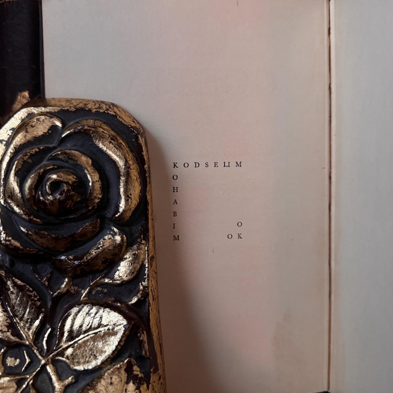 Op de afbeelding: Een close-up van een boek met een gouden bloemenontwerp op de rug, tegen een zwarte achtergrond. De open pagina toont de tekst "KODSELIM O HARIM O OK".