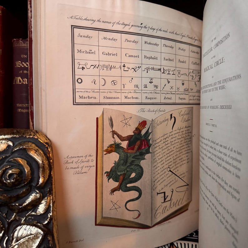 Puede incluir: Un libro antiguo abierto con ilustraciones detalladas y texto manuscrito. Las p&aacute;ginas muestran una tabla de &aacute;ngeles y un dibujo colorido de una figura montando un drag&oacute;n. El t&iacute;tulo del libro es "The Book of Spirits".