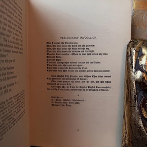 Op de afbeelding: Open boek met de tekst "PRELIMINARY INVOCATION" in zwarte letters op verouderd papier. De boekband, met gouden en zwarte versieringen, is rechts zichtbaar. Het paginanummer "vii" staat onderaan.