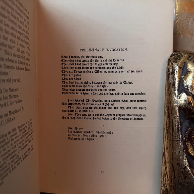 Op de afbeelding: Open boek met de tekst "PRELIMINARY INVOCATION" in zwarte letters op verouderd papier. De boekband, met gouden en zwarte versieringen, is rechts zichtbaar. Het paginanummer "vii" staat onderaan.