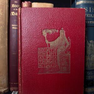 May include: A red book with gold lettering and a detailed illustration on the cover. The cover text includes the words "DEATH," "ALPHA," and "HTAED." The book is part of a collection of antique books.