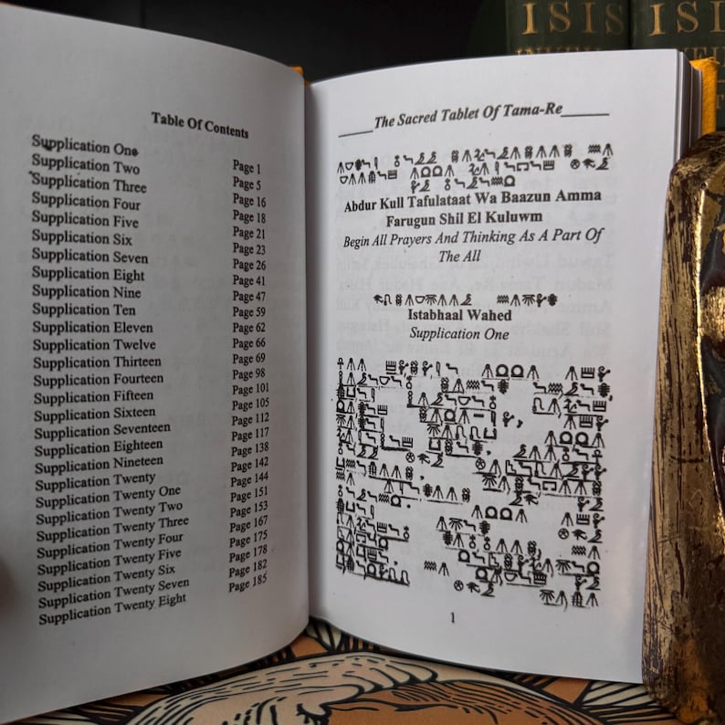 May include: An open book with text and hieroglyphs on its pages. The left page displays a table of contents, and the right page features ancient script and the title "The Sacred Tablet of Tama-Re". The book is open to page one.