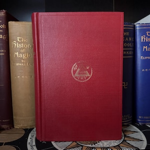May include: A red book with gold lettering and a gold emblem. The book is titled "TRY" and is part of a collection of books on a shelf. Other books include "The History of Magic" and "The Cane Tools".