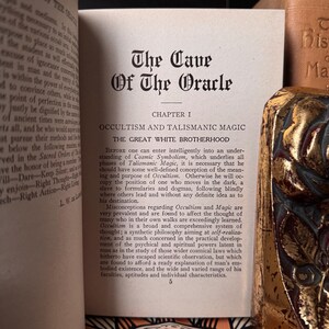 May include: Open book with the title "The Cave of the Oracle" and the chapter heading "Occultism and Talismanic Magic." The pages are aged, with text in black font. A decorative gold book is visible on the right side.