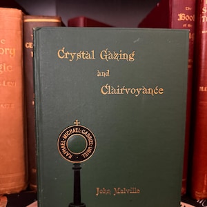 Könnte beinhalten: Ein grünes Buch mit dem Titel "Crystal Gazing and Clairvoyance" von John Melville. Der Titel ist in goldener Schrift gehalten. Ein kreisförmiges Emblem mit den Worten "Raphael Michael Gabriel Uriel" umgibt ein Kristallkugelbild. Andere Bücher sind im Hintergrund sichtbar.