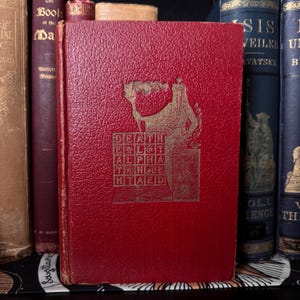 May include: A vintage red book with gold lettering and a detailed illustration on the cover. The cover text includes the words "DEATH," "ALPHA," and "HTAED." The book is surrounded by other antique books, suggesting a collection of historical literature.