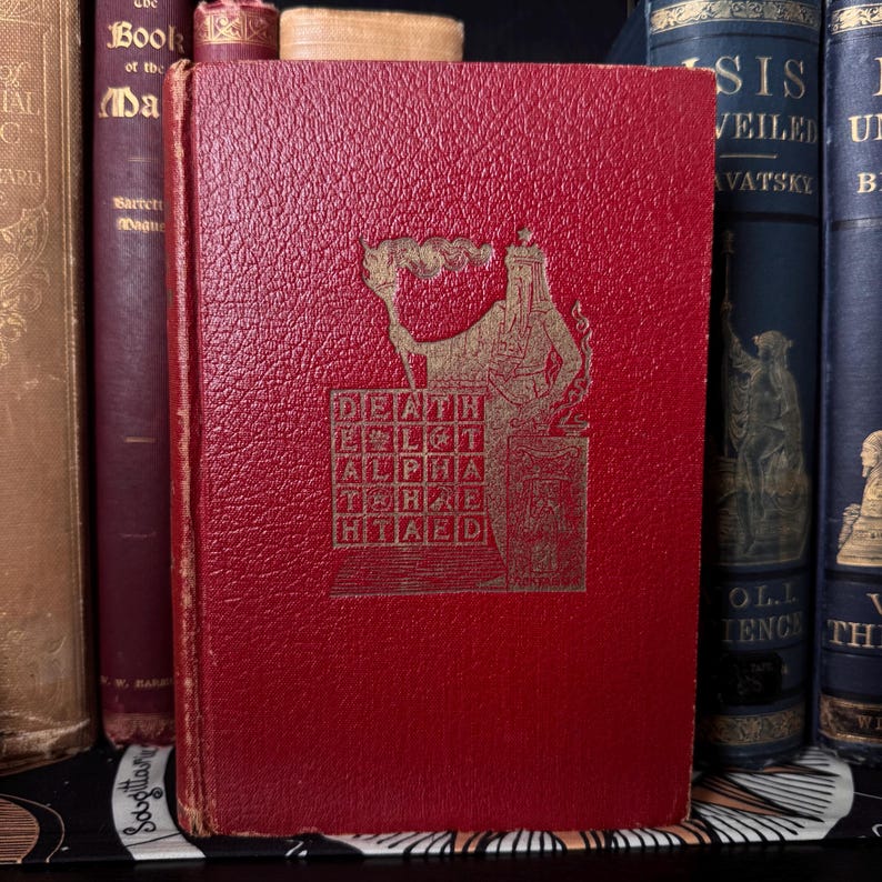 May include: A vintage red book with gold lettering and a detailed illustration on the cover. The cover text includes the words "DEATH," "ALPHA," and "HTAED." The book is surrounded by other antique books, suggesting a collection of historical literature.