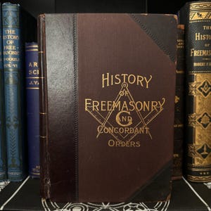 Op de afbeelding: Een bruin lederen gebonden boek met gouden letters met de titel "Geschiedenis van de Vrijmetselarij en de Concordante Orden". De omslag toont een gouden vrijmetselaars symbool.
