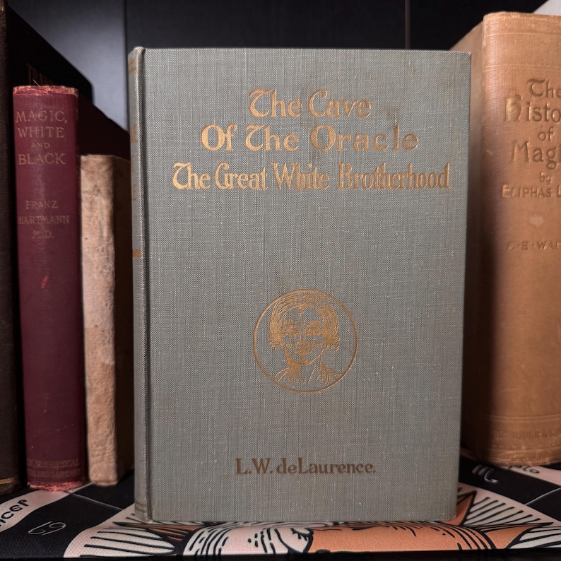 May include: A vintage book titled "The Cave of The Oracle The Great White Brotherhood" with gold lettering on a gray-green cover. The cover features a circular illustration and the author's name, L.W. deLaurence. Other antique books are visible in the background.