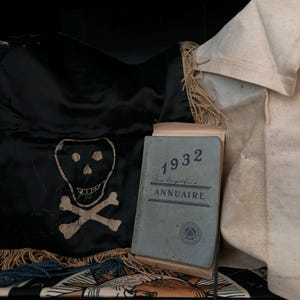 May include: A black fabric banner with a white skull and crossbones design. A blue and gold fringed trim surrounds the banner. A blue book with the text "1932 Annuaire" is in the foreground.