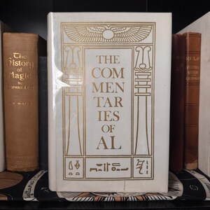 May include: A white book titled "The Commentaries of Al" with gold lettering and Egyptian-style illustrations. The book is flanked by other antique books with brown covers, suggesting a collection of historical or esoteric texts.