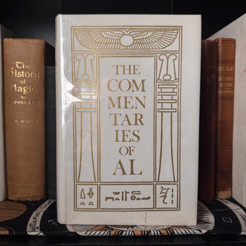 May include: A white book titled "The Commentaries of Al" with gold lettering and Egyptian-style illustrations. The book is flanked by other antique books with brown covers, suggesting a collection of historical or esoteric texts.